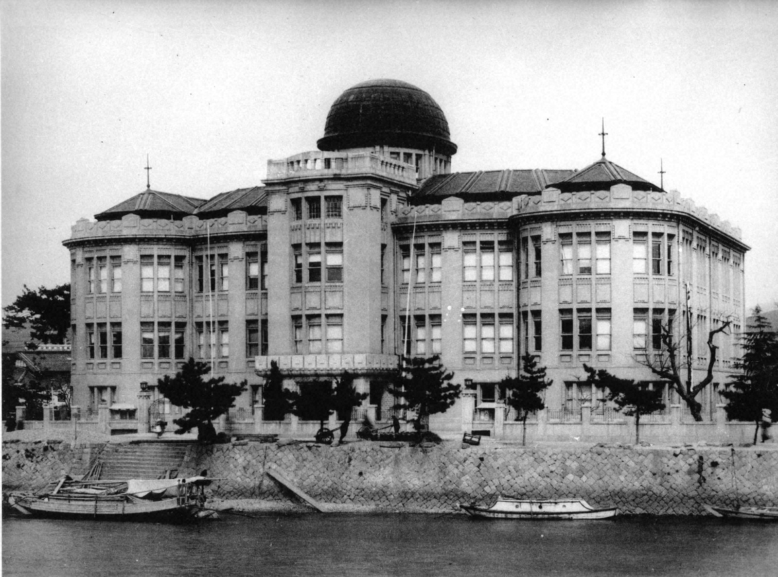A photo of the Hiroshima Prefectural Commercial Exhibition Hall before the bomb. The building has three stories and what looks like two wings to it. There is a rounded dome positioned in the middle of its roof. The shot has been taken from the river to the back of the building and there are small boats visible in the lower part of the photo.  