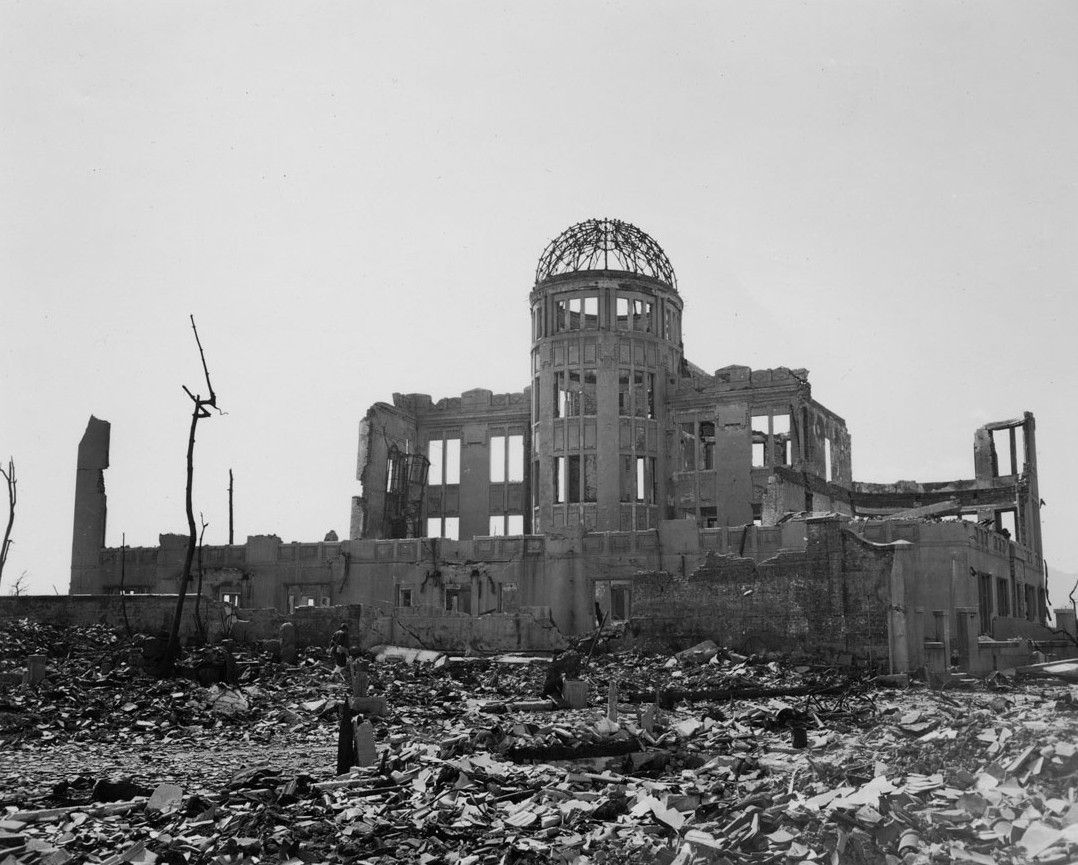 A photo showing Hiroshima Prefectural Commercial Exhibition Hall after the blast. The exterior walls have all been destroyed along with the roof and upper levels. All that remains is the foundation walls of the ground floor and the skeletal frame of the cylindrical structure holding up the dome, which itself has been reduced to a wire frame. The sounding area is covered in layers of rubble and the trees in front of the building are nothing more than chard sticks.