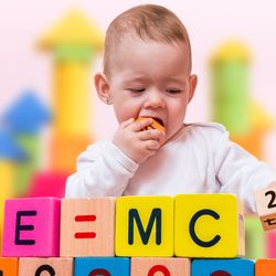 A photo of a baby plying with wooden bricks that spell out "E=MC2". The Child is placing the 2 brick on the end of the sequence and has another brick in its hand which it is about to chew on. Little genius. 