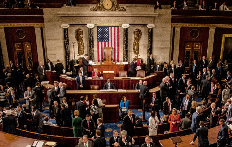 A photo of members of congress mingling on the house floor. The photo captures dozens of people talking to one another and the American flag is situated in the center of the image.