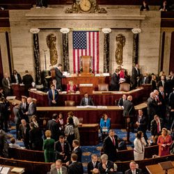 A photo of members of congress mingling on the house floor. The photo captures dozens of people talking to one another and the American flag is situated in the center of the image.