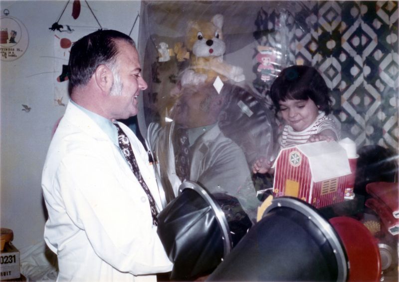 David Vetter, a young boy with dark hair, pictured inside the plastic sterile "bubble" in which he lived. Immunologist John R. Montgomery interacts with David via a glovebox.