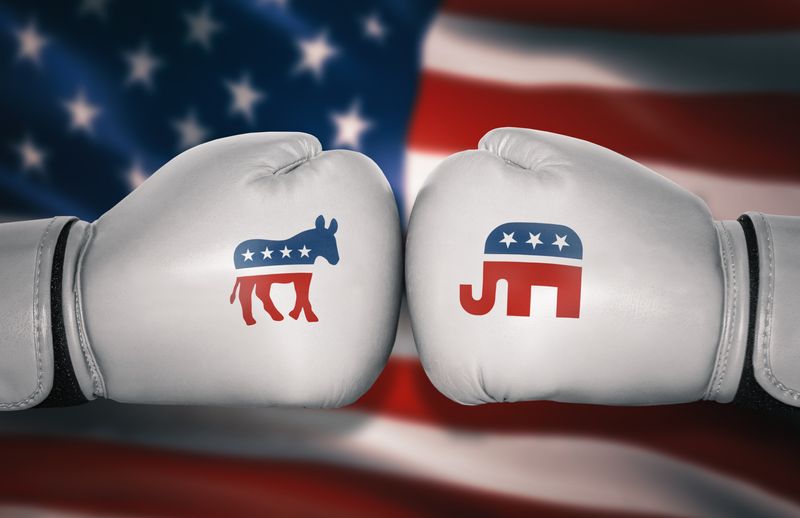 A photo shows two white boxing gloves pressed against one another. On the left is the symbol of the Democratic party as a donkey, while the glove on the right has an elephant for the Republicans. The two gloves are positioned in front of an American flag. 