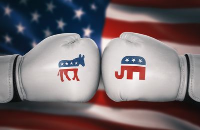 A photo shows two white boxing gloves pressed against one another. On the left is the symbol of the Democratic party as a donkey, while the glove on the right has an elephant for the Republicans. The two gloves are positioned in front of an American flag. 