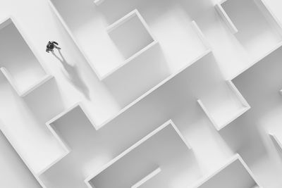A photo of a male figure walking through a white maze. The image is looking down on the figure from above. His shadow is being cast in front of him as he walks through this white space.
