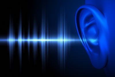 A digital image showing a close up of a human ear on the far right. There are sound waves represented as spiky lines of light entering the ear. Both the ear and the light consist of blue tones and both are set against a black background. 