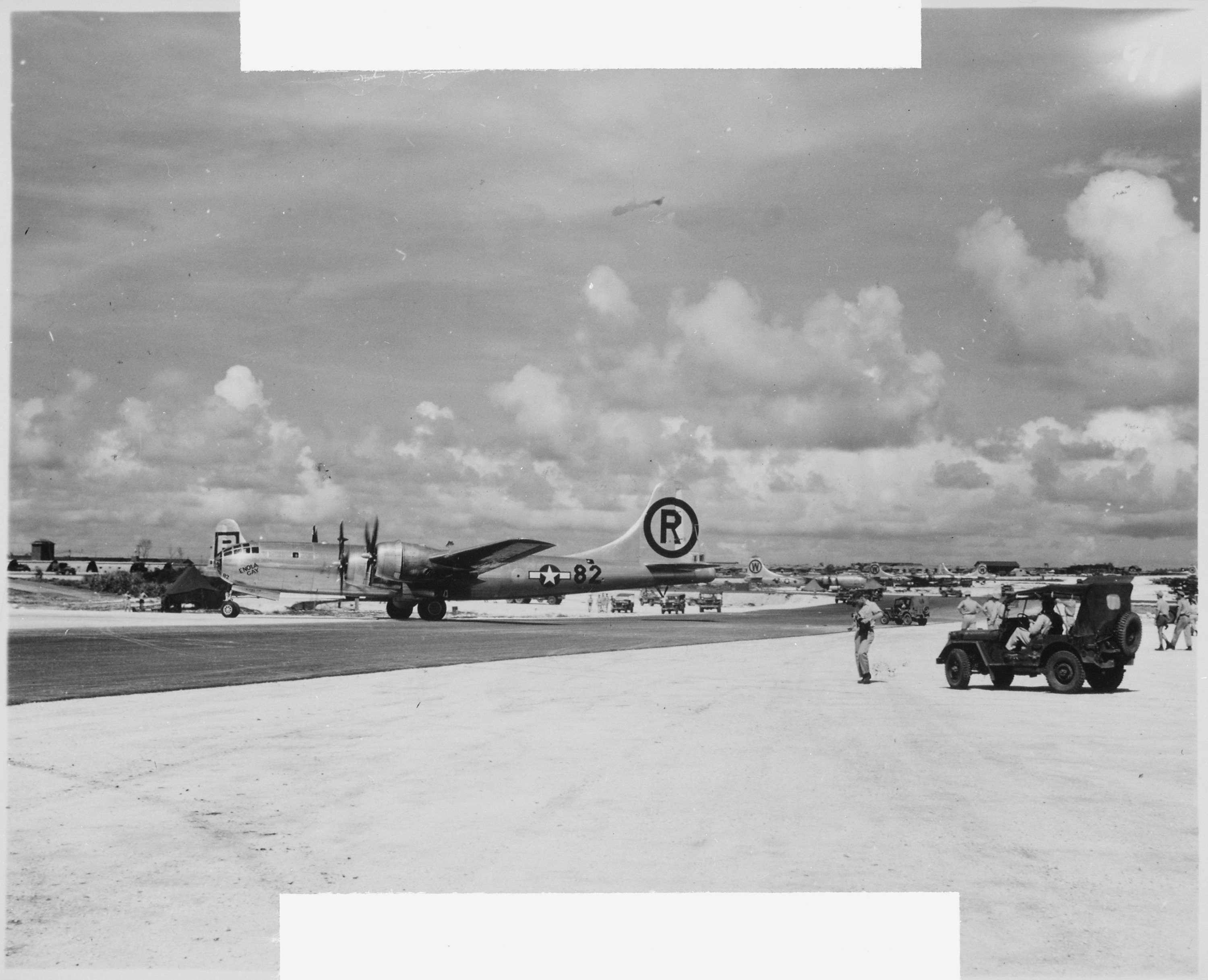 A black and white photo showing the Enola Gay on a runway. The aircraft is shown on profile, facing the left of the shot. It has twin predellas on each wings. There is a large R encircled and printed on its tail and the number 82 is printed on its tail, next to a symbol of a star in a circle with a rectangle running through it. The word "Enola Gay" is written at an angle just under the cockpit window. The aircraft is in the middle of the shot. In the righthand side of the foreground is a jeep with several military personnel standing around it. 