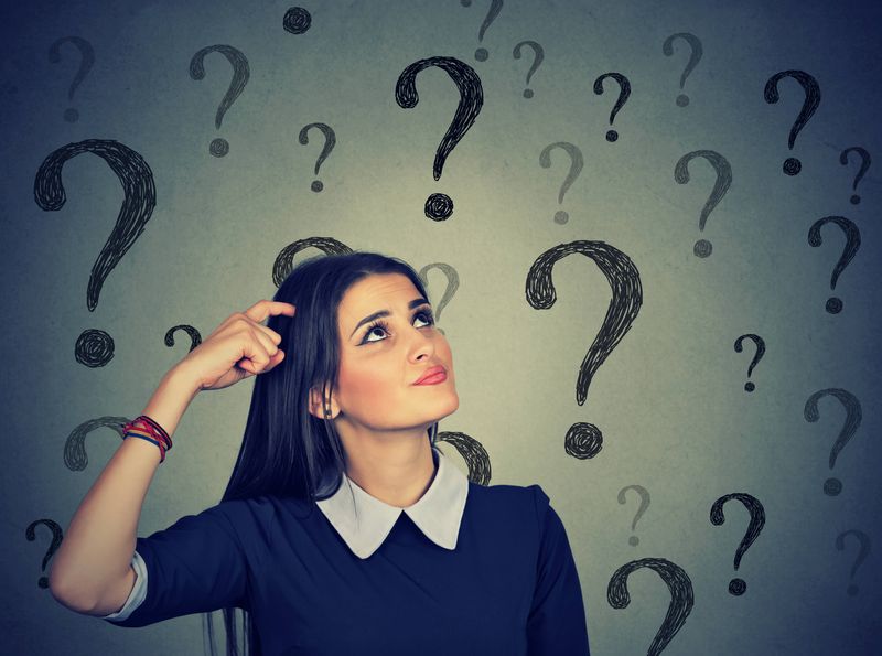 The photo shows a young woman wearing a dark top with a tick white collar looking up and scratching her head. She has a slightly confused look on her face. There are large question marks floating around her. 