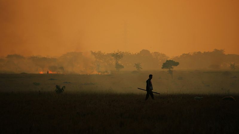 Silhouette of a man walking across a hazy field with a wildfire in background.