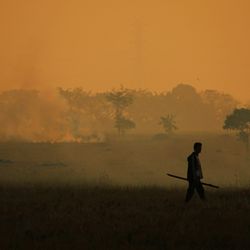 Silhouette of a man walking across a hazy field with a wildfire in background.