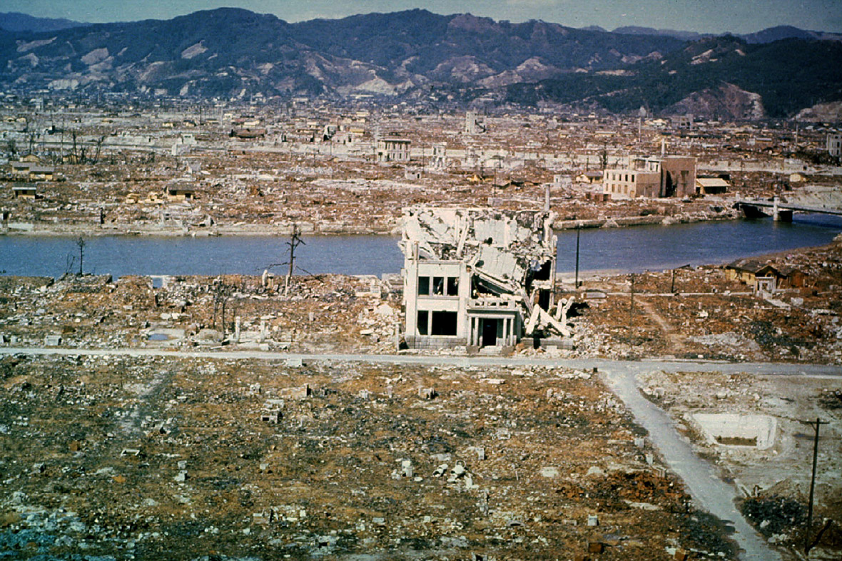 A colored photo showing Hiroshima after the explosion. A few concrete structures are still standing, though they are mostly rubble. The rest of the shot shows the city flattened by the blast. The shot extends all the way to the mountains in the distance. 