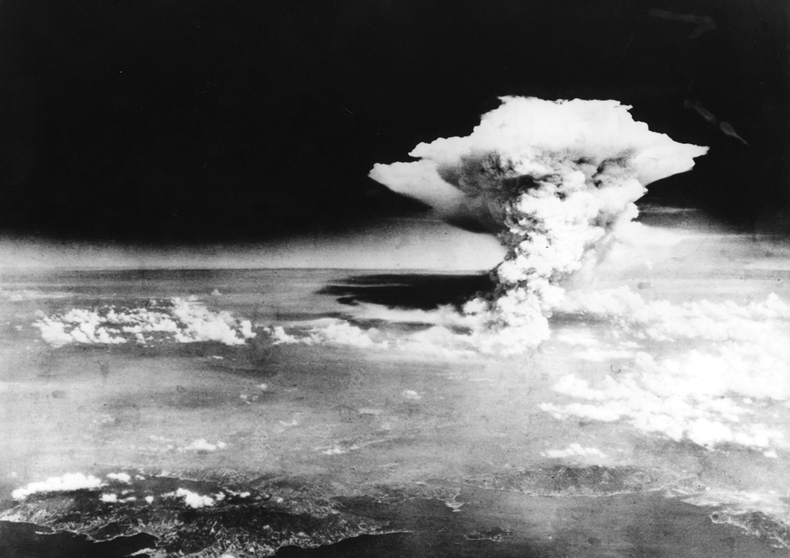 A photo taken from the Necessary Evil showing the mushroom cloud towering above the city of Hiroshima. The cloud occupies the center left of the shot and there is denser cloud covering the lower portion of the photos. However, in the bottom part of the photo, you can see the coast line which provides scale for how enormous the cloud is.