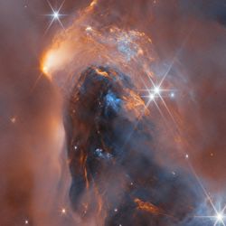 A nebula made up of cloudy gas and dust in the form of soft and wispy clouds and, in the centre, thin and highly detailed layers pressed close together. Large, bright stars surrounded by six long points of light are dotted over the image, as well as some small, point-like stars embedded in the clouds. The clouds are lit up in blue close to the stars; orange colours show clouds that glow in infrared light.