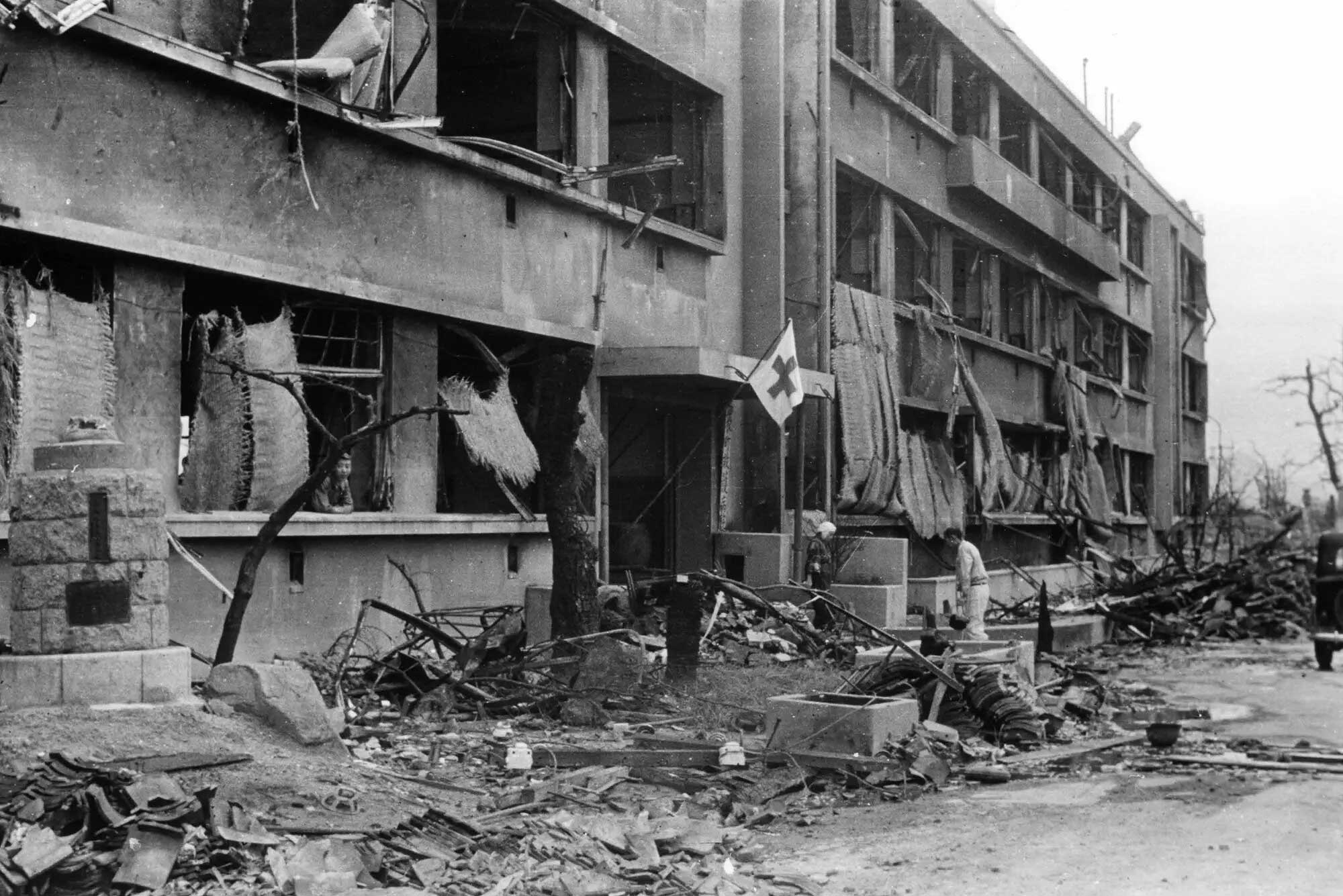 The photo shows a lopsided Red Cross flag attached to the entrance of an otherwise blown out three storied building. The windows are all missing and the front of the building is strewn with rubble and twisted metal. There is a man shown walking towards the entrance. 