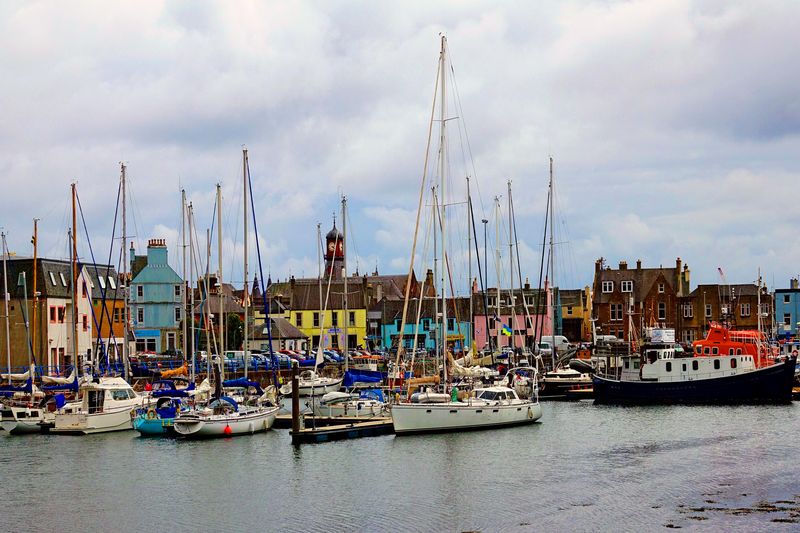 The photo shows a dock at low tide with multiple small boats and dinghies. In the background, the buildings of a town can be seen huddled around the dock. The building in the centre of this mass has a yellow face, while behind it the top of a church is visible. The buildings next to it have pink and blue faces while others are red brick.