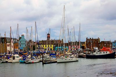 The photo shows a dock at low tide with multiple small boats and dinghies. In the background, the buildings of a town can be seen huddled around the dock. The building in the centre of this mass has a yellow face, while behind it the top of a church is visible. The buildings next to it have pink and blue faces while others are red brick.