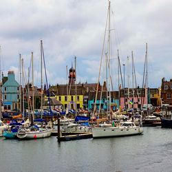 The photo shows a dock at low tide with multiple small boats and dinghies. In the background, the buildings of a town can be seen huddled around the dock. The building in the centre of this mass has a yellow face, while behind it the top of a church is visible. The buildings next to it have pink and blue faces while others are red brick.