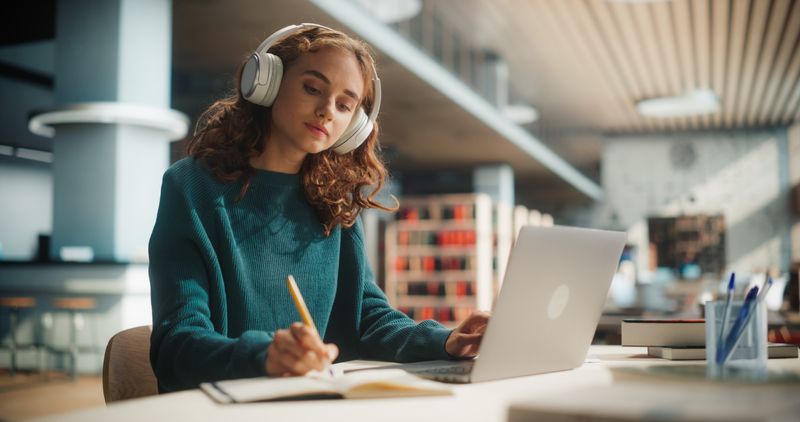 The photo shows a young woman sitting in a university library as she listens to something in front of her laptop. She has large white headphones on and is writing something in a note book. 