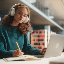 The photo shows a young woman sitting in a university library as she listens to something in front of her laptop. She has large white headphones on and is writing something in a note book. 
