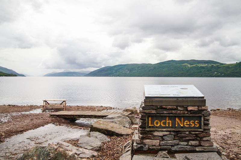 A photo taken on the shore of Loch Ness on a grey cloudy day. In the foreground a stone monument is visible with the words "Lock Ness" on it. It has a square metal plaque on top. The beach leading down to the water is covered in shingle stone and then there is a flat body of water stretching off. In the distance, a deep green landmass is visible set against low hanging clouds.
