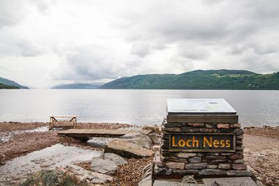 A photo taken on the shore of Loch Ness on a grey cloudy day. In the foreground a stone monument is visible with the words "Lock Ness" on it. It has a square metal plaque on top. The beach leading down to the water is covered in shingle stone and then there is a flat body of water stretching off. In the distance, a deep green landmass is visible set against low hanging clouds.