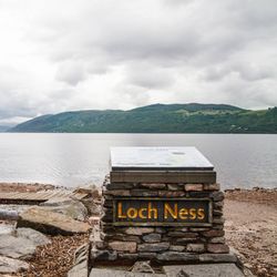 A photo taken on the shore of Loch Ness on a grey cloudy day. In the foreground a stone monument is visible with the words "Lock Ness" on it. It has a square metal plaque on top. The beach leading down to the water is covered in shingle stone and then there is a flat body of water stretching off. In the distance, a deep green landmass is visible set against low hanging clouds.