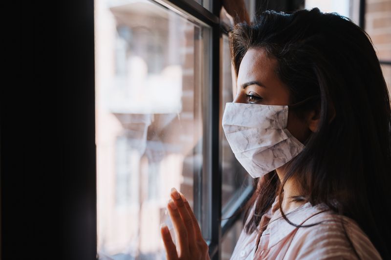 woman with long dark hair wearing a white homemade fabric face covering, looking towards the left out of a window with one hand pressed against the glass