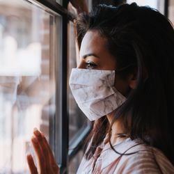 woman with long dark hair wearing a white homemade fabric face covering, looking towards the left out of a window with one hand pressed against the glass