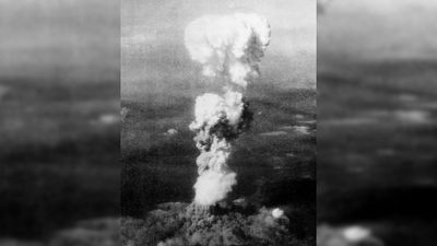 An aerial photo of the mushroom cloud looming over Hiroshima. It is a large thin stalk of cloud with the top starting to bud into the classic shape. 