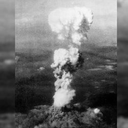 An aerial photo of the mushroom cloud looming over Hiroshima. It is a large thin stalk of cloud with the top starting to bud into the classic shape. 