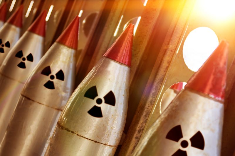 A photo showing a line of nuclear warheads attached to missiles. They are the pointed red tips on five weapons that have the radiation symbol just under them. The image shows the tops of these missiles lined up against a metal stand that has circular gaps in it which are letting in light.