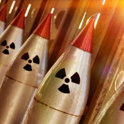 A photo showing a line of nuclear warheads attached to missiles. They are the pointed red tips on five weapons that have the radiation symbol just under them. The image shows the tops of these missiles lined up against a metal stand that has circular gaps in it which are letting in light.