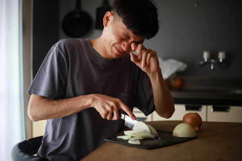 A photo shows a man at a kitchen counter cutting an onion. His face is scrunched up and he is rubbing his left eye with his left hand. 