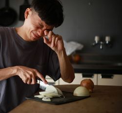 A photo shows a man at a kitchen counter cutting an onion. His face is scrunched up and he is rubbing his left eye with his left hand. 