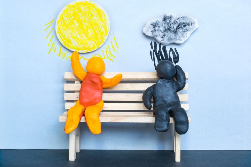 Concept of optimism and pessimism. Two clay figures sitting on a bench. The one on the right is composed of orange and red clay and has a sun above them, while the one on the left is composed of deep blue/black clay and has a raincloud above them.