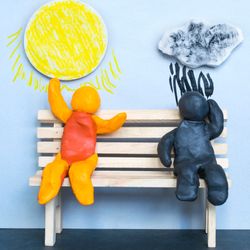Concept of optimism and pessimism. Two clay figures sitting on a bench. The one on the right is composed of orange and red clay and has a sun above them, while the one on the left is composed of deep blue/black clay and has a raincloud above them.
