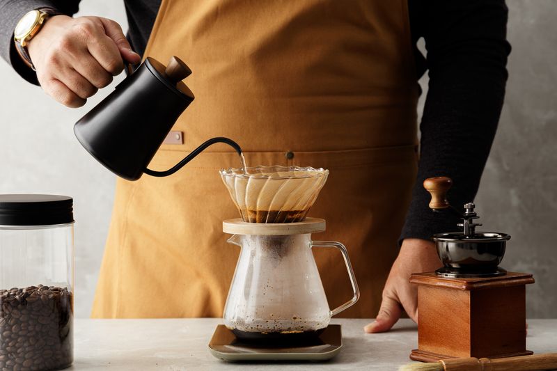 barista making pour-over coffee using a gooseneck kettle, with a grinder and a jar of beans on the table next to the pot