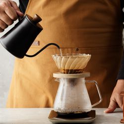 barista making pour-over coffee using a gooseneck kettle, with a grinder and a jar of beans on the table next to the pot
