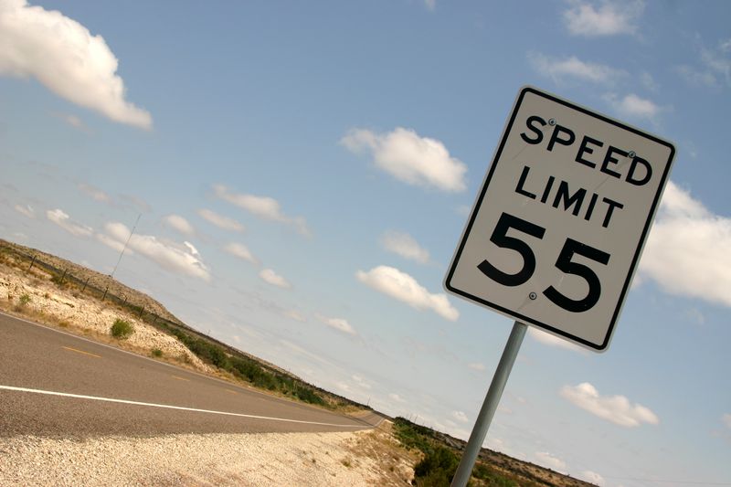 A photo of a "Speed Limit" sign on the side of a road. The signs in a white rectangle with the words "Speed Limit 55" written on it. The photo shows the sign  on the far right of the image, with the road to the left. The image is framed at an angle and the road appears to be passing through a desert. 