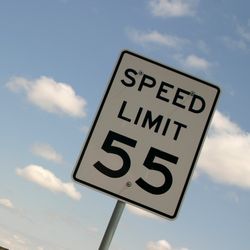 A photo of a "Speed Limit" sign on the side of a road. The signs in a white rectangle with the words "Speed Limit 55" written on it. The photo shows the sign  on the far right of the image, with the road to the left. The image is framed at an angle and the road appears to be passing through a desert. 