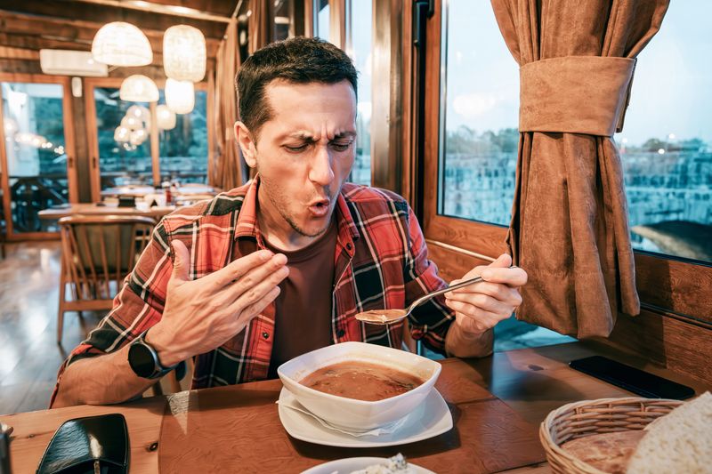 man eating spicy soup wearing a pained expression and wafting his face in the heat. He is in a restaurant with lots of wood panelling sitting next to a window, wearing a red plaid shirt