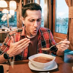man eating spicy soup wearing a pained expression and wafting his face in the heat. He is in a restaurant with lots of wood panelling sitting next to a window, wearing a red plaid shirt