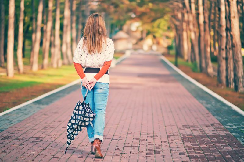 woman viewed from behind strolling along a park path lined with trees. She has long dark blond hair and is wearing a cream sweater over an orange long-sleeve shirt, a brown belt, jeans, and tan boots. She is carrying a black and white checked umbrella behind her back