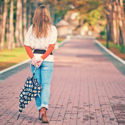 woman viewed from behind strolling along a park path lined with trees. She has long dark blond hair and is wearing a cream sweater over an orange long-sleeve shirt, a brown belt, jeans, and tan boots. She is carrying a black and white checked umbrella behind her back