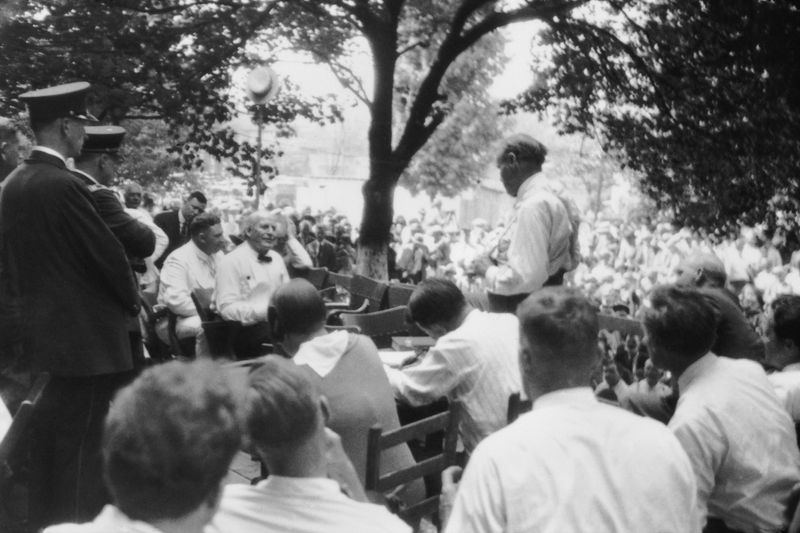 An old black and white photo showing Clarence Darrow interrogating William Jennings Bryan outside under some trees. Darrow is standing with his back to the camera while Bryan is sat opposite him. The two men are surrounded by dozens of seated people listening to the trial.