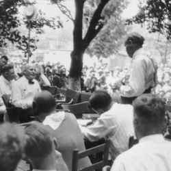 An old black and white photo showing Clarence Darrow interrogating William Jennings Bryan outside under some trees. Darrow is standing with his back to the camera while Bryan is sat opposite him. The two men are surrounded by dozens of seated people listening to the trial.