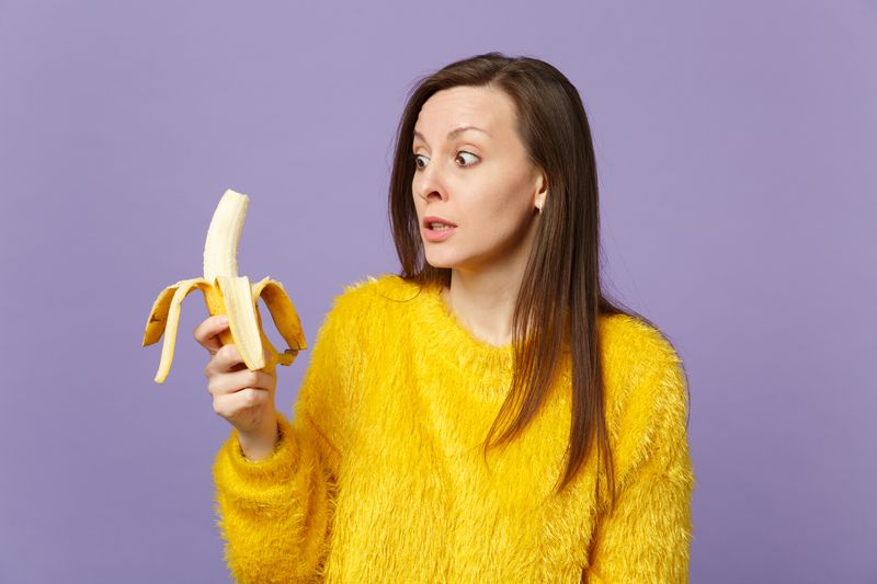 woman in yellow sweater looking slightly frightened at a half-peeled banana in her hand, on a lilac background