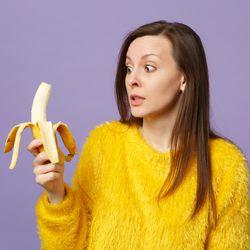woman in yellow sweater looking slightly frightened at a half-peeled banana in her hand, on a lilac background