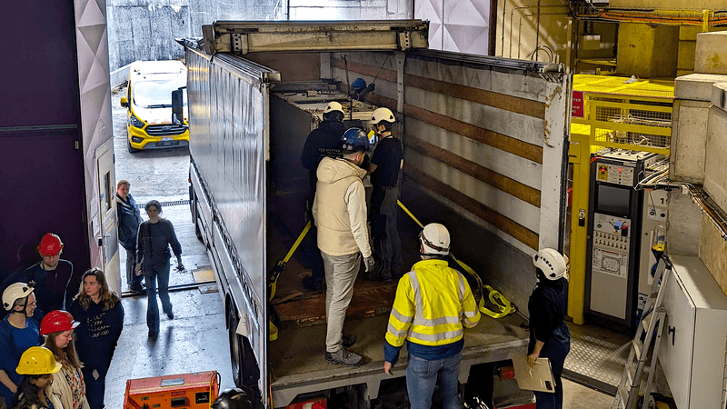 an open van is holding a metallic cuboid. several people are attaching ropes to keep it stable.
