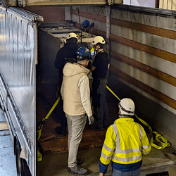 an open van is holding a metallic cuboid. several people are attaching ropes to keep it stable.